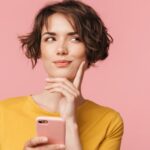 Thinking dreaming young beautiful woman posing isolated over pink wall background using mobile phone.